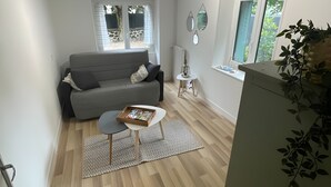 A compact living space with a grey sofa, a small round table, a radiator, and a window with a view of the outdoors. - Le Puy d’Eraigne: peaceful, cool apartment with terrace and optional garage on request (Saint-Nectaire)