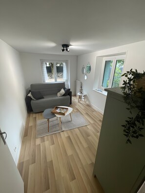 Un espace de vie compact comprenant un canapé gris, une petite table ronde, un radiateur et une fenêtre donnant sur l’extérieur.