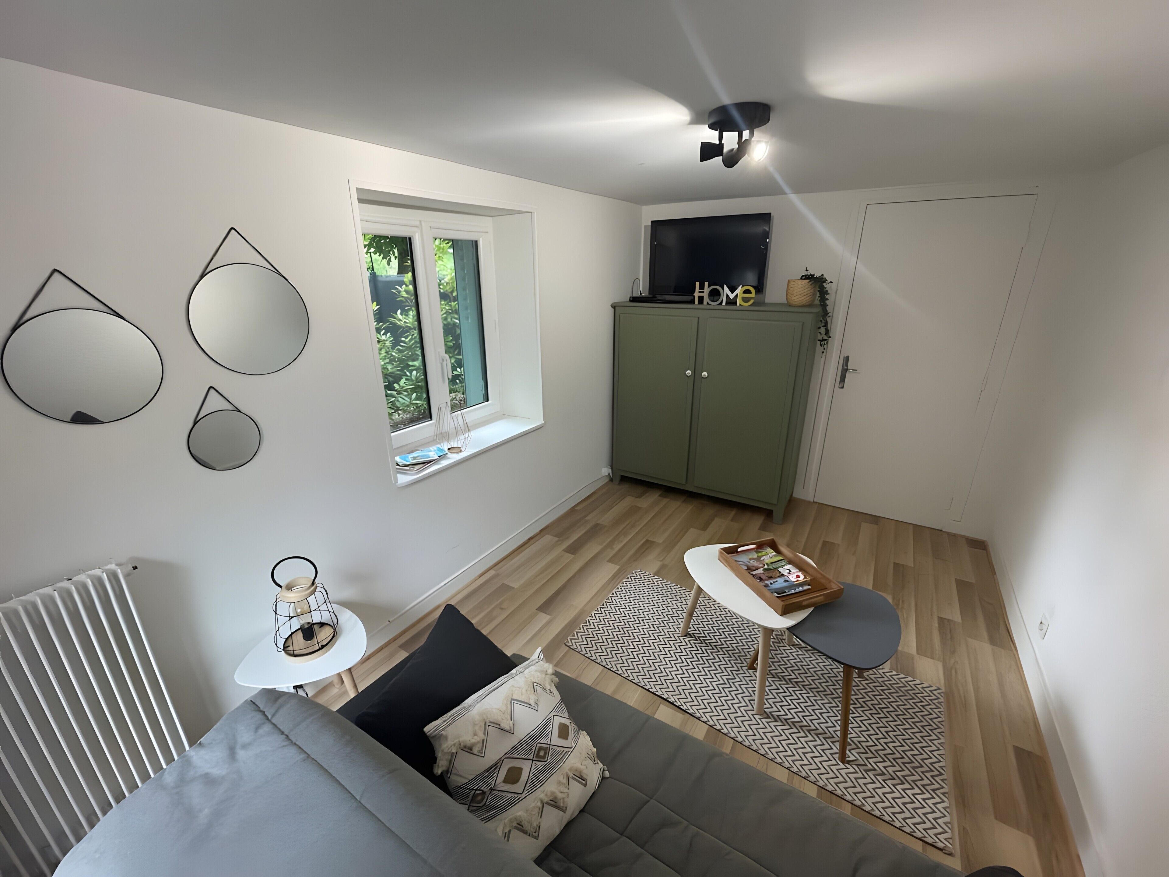 A modern living room with a grey sofa, a small white table, a green cabinet, and a window with a view of greenery.