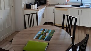 A kitchen with white cabinets, a dining table with chairs, a green folder, and a window with a view of greenery. - Le Puy d’Eraigne: peaceful, cool apartment with terrace and optional garage on request (Saint-Nectaire)