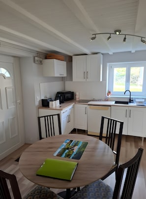 A kitchen with white cabinets, a dining table with chairs, a green folder, and a window with a view of greenery. - Le Puy d’Eraigne: peaceful, cool apartment with terrace and optional garage on request (Saint-Nectaire)