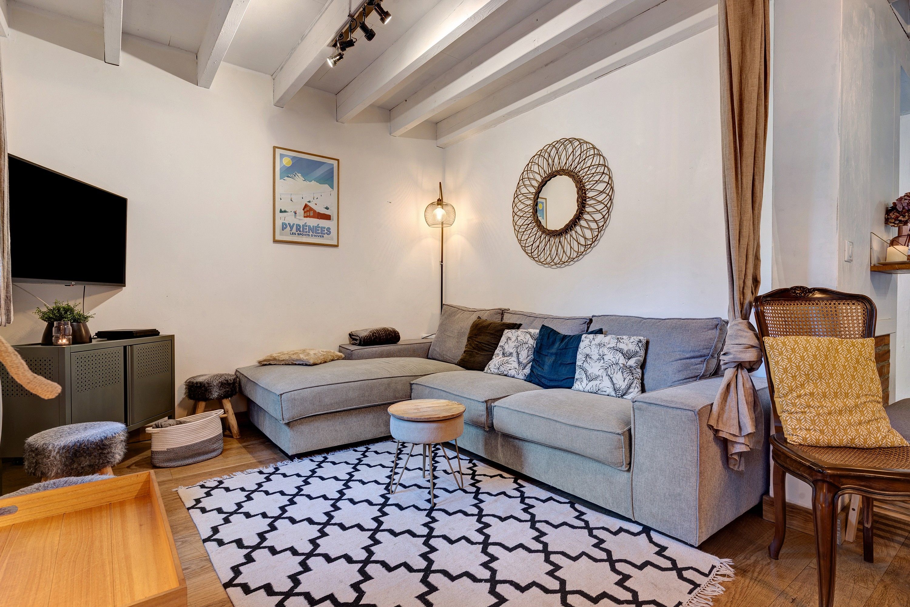 Een moderne woonkamer met een grijze modulaire bank, een getextureerd vloerkleed, een houten salontafel, een rieten fauteuil en een flatscreen tv aan de muur.