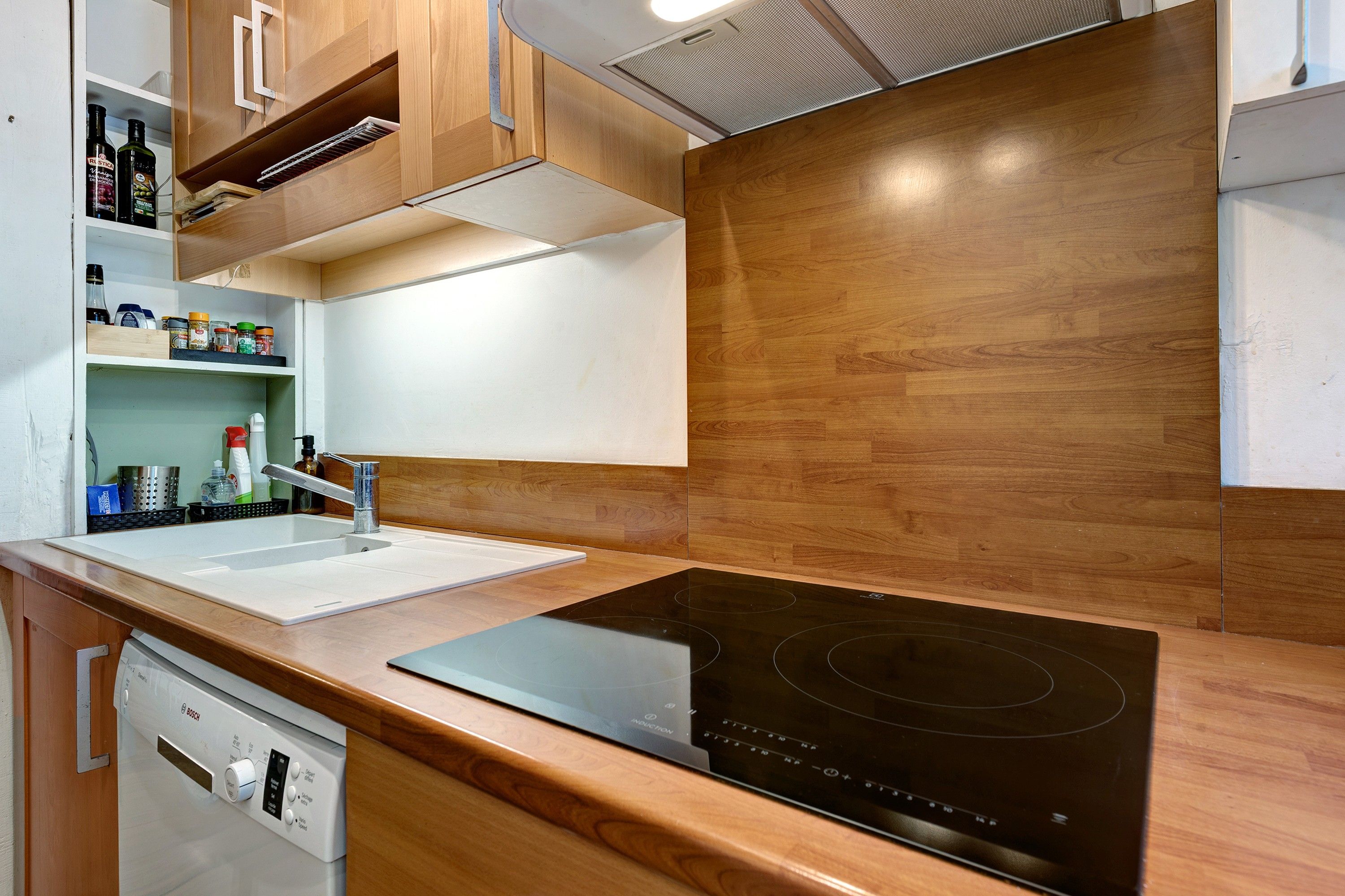 Une cuisine moderne équipée d’un lave-vaisselle Bosch, d’une plaque de cuisson à induction noire, de meubles en bois et d’un plan de travail blanc.