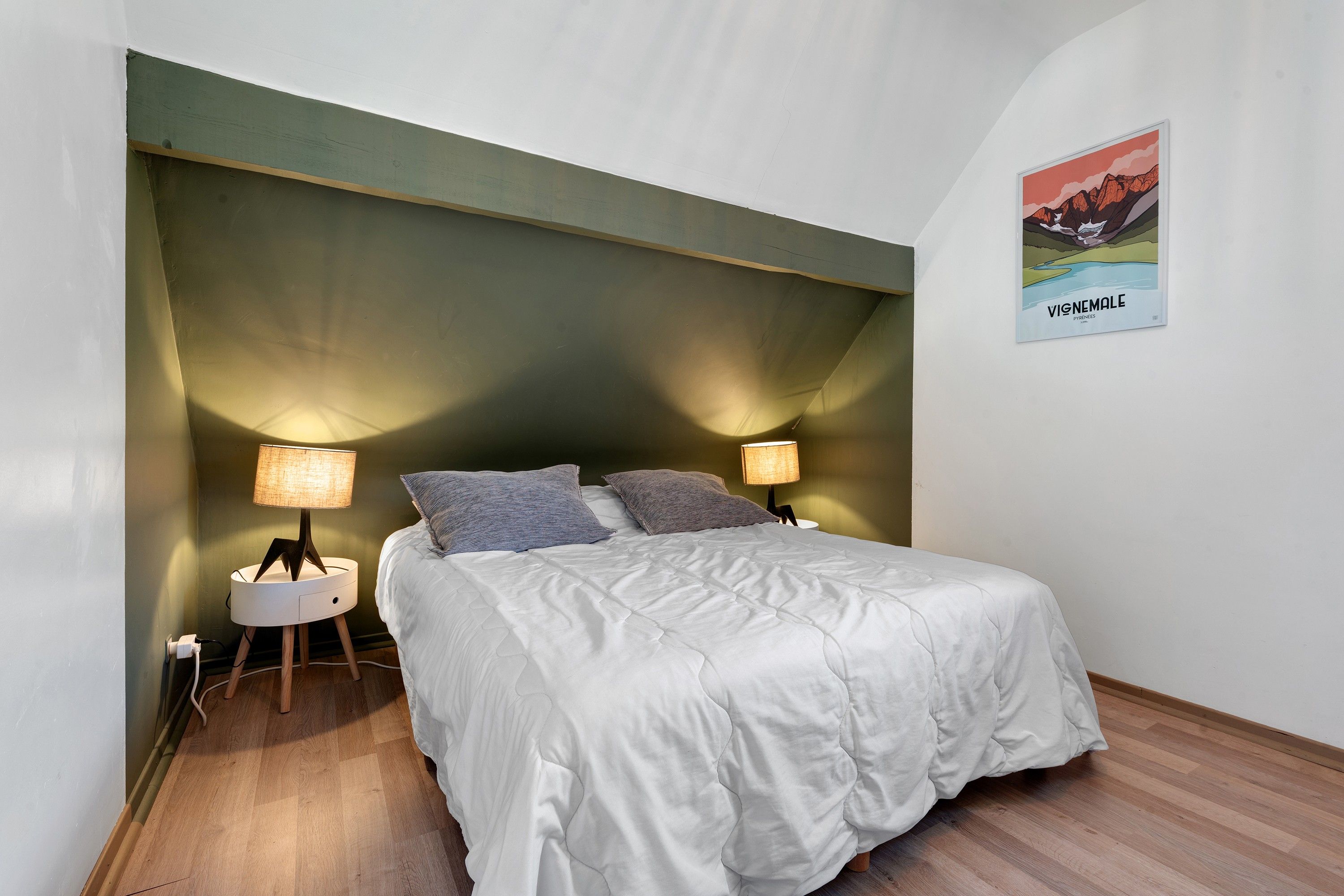 Een slaapkamer met een bed, twee bedlampen, een nachtkastje, een aan de muur hangend schilderij en een houten vloer.