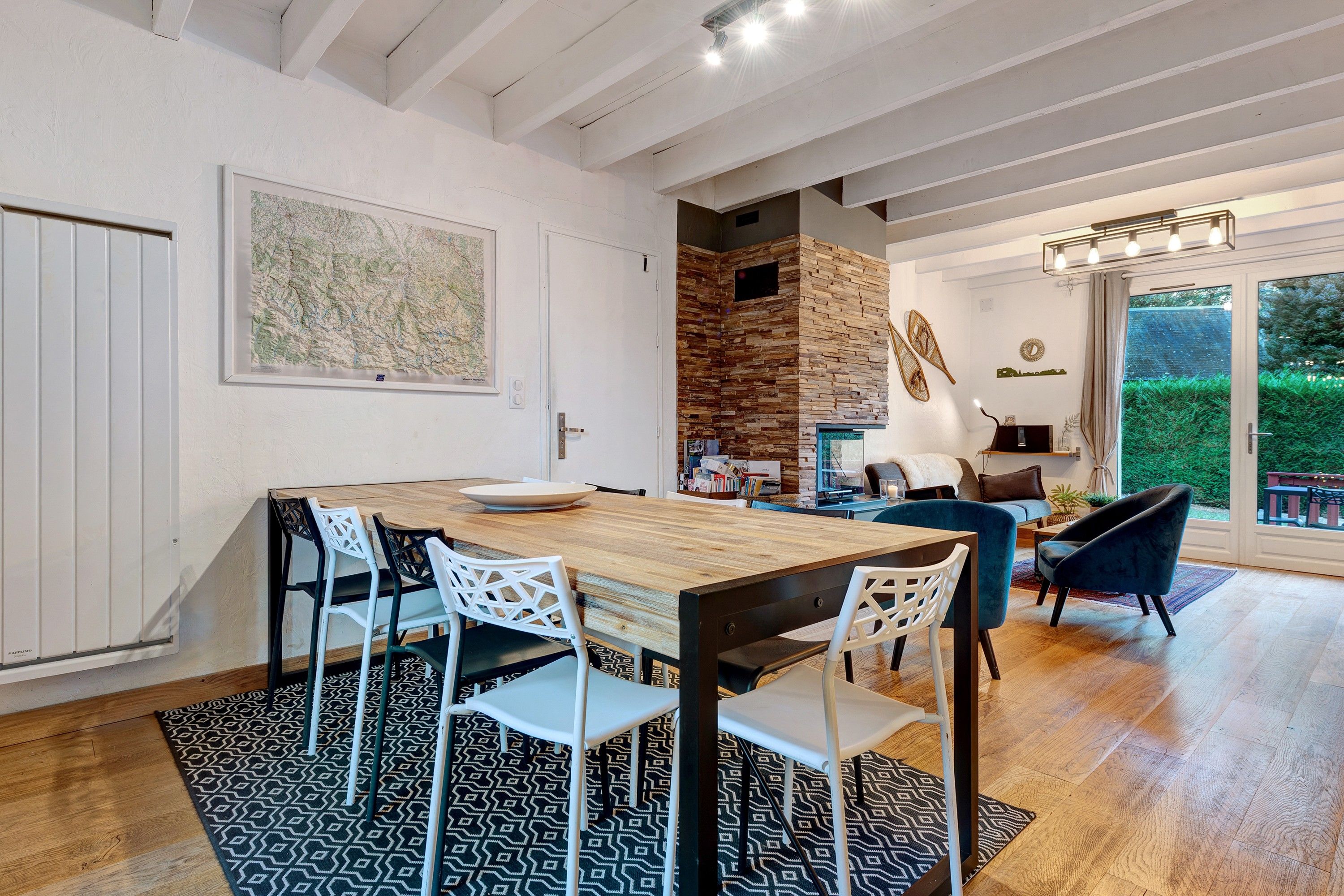 Een eetgedeelte met een houten tafel en stoelen, een keuken met een stenen muur, en uitzicht op een tuin door een glazen deur.