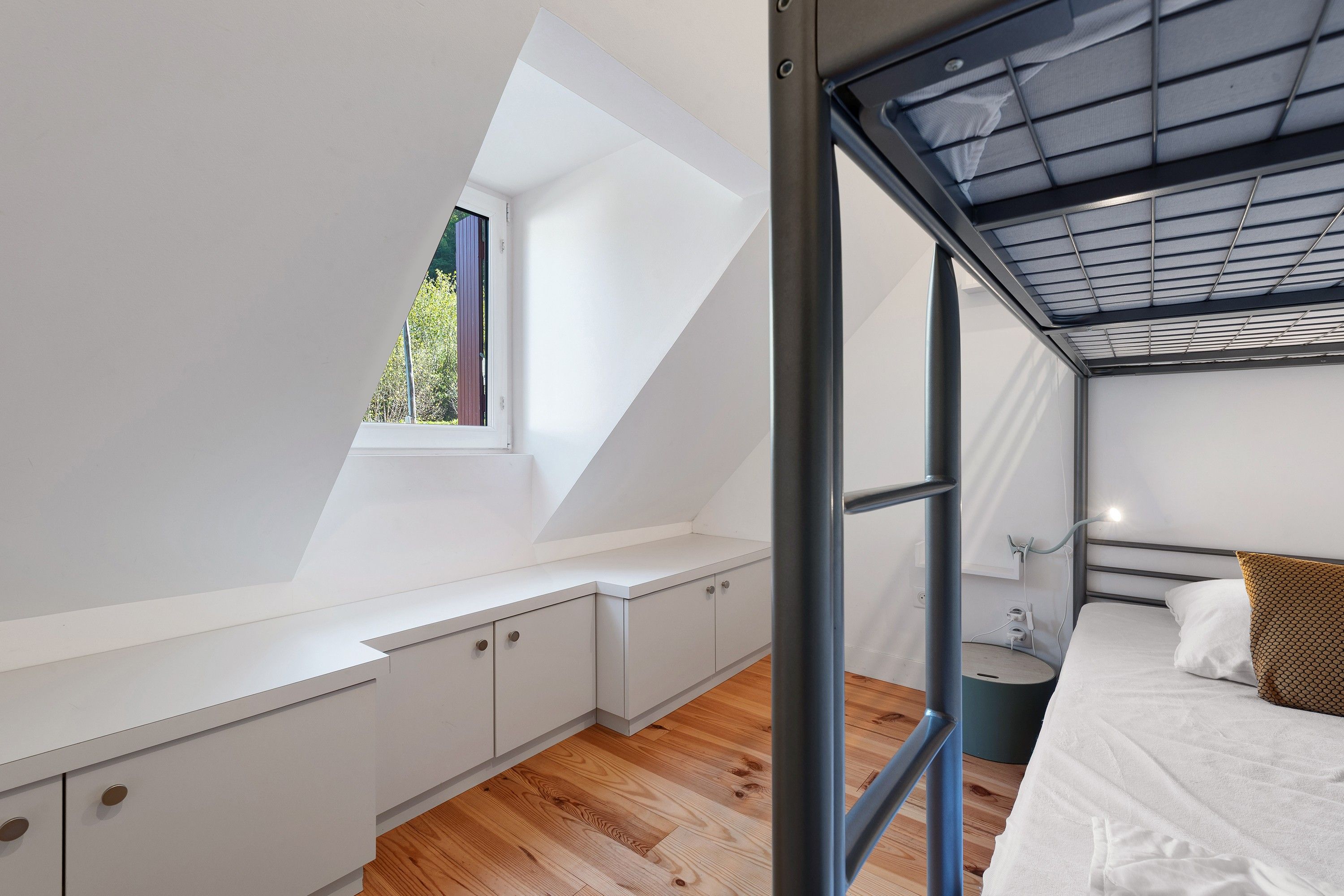 Una habitación moderna y compacta con techo inclinado, una cama litera, un ventanal pequeño y un escritorio empotrado.