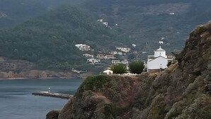 Paisagem costeira com terreno rochoso, um farol e edifícios numa encosta.