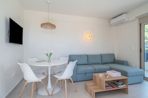 Ein modernes Wohnzimmer mit grauem Sofa, weißen Sesseln, einem runden Esstisch, einer Pendelleuchte und einem an der Wand befestigten Flachbildfernseher.