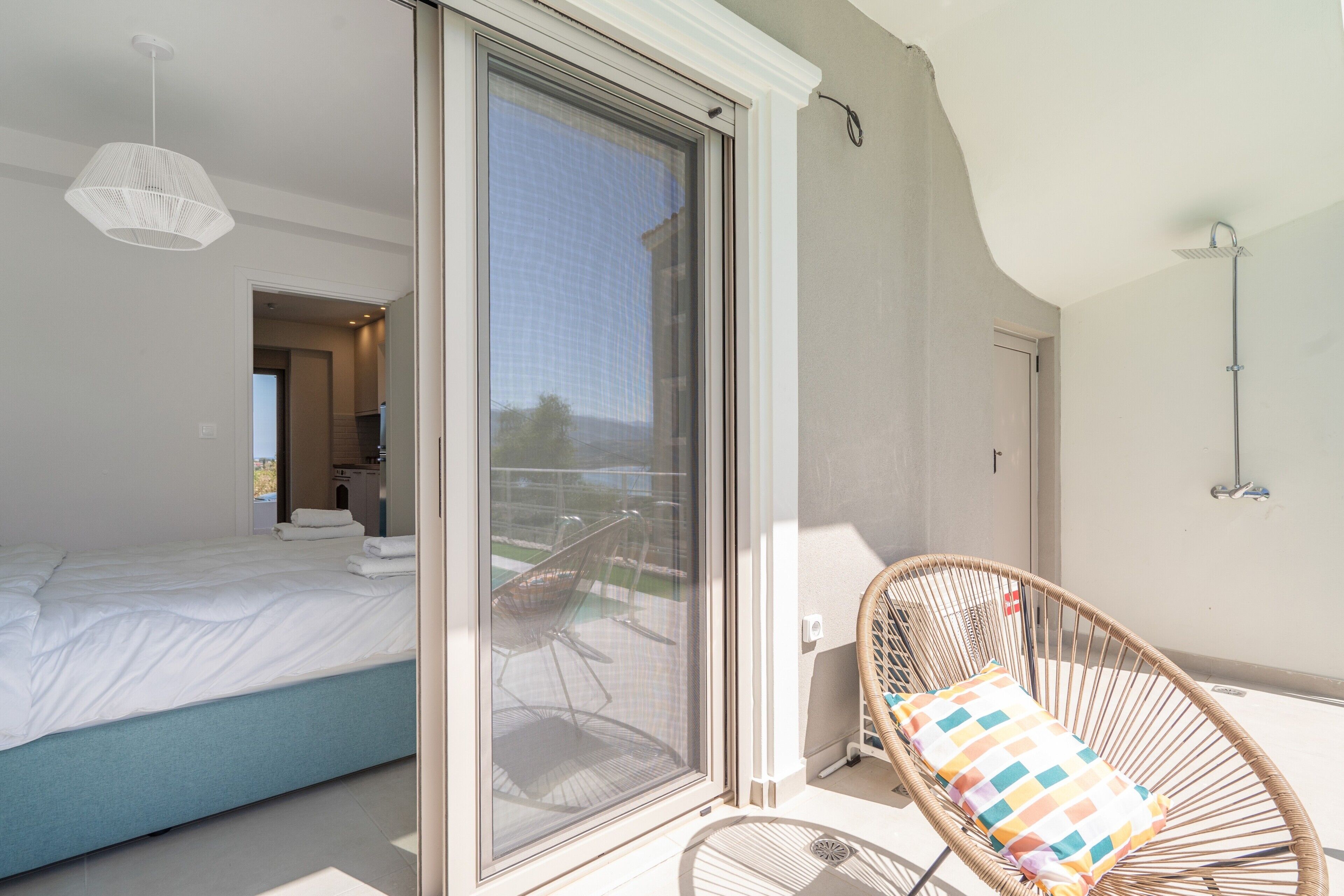 A bedroom with a bed, a wicker chair with a cushion, a sliding glass door, and a view of the outside.
