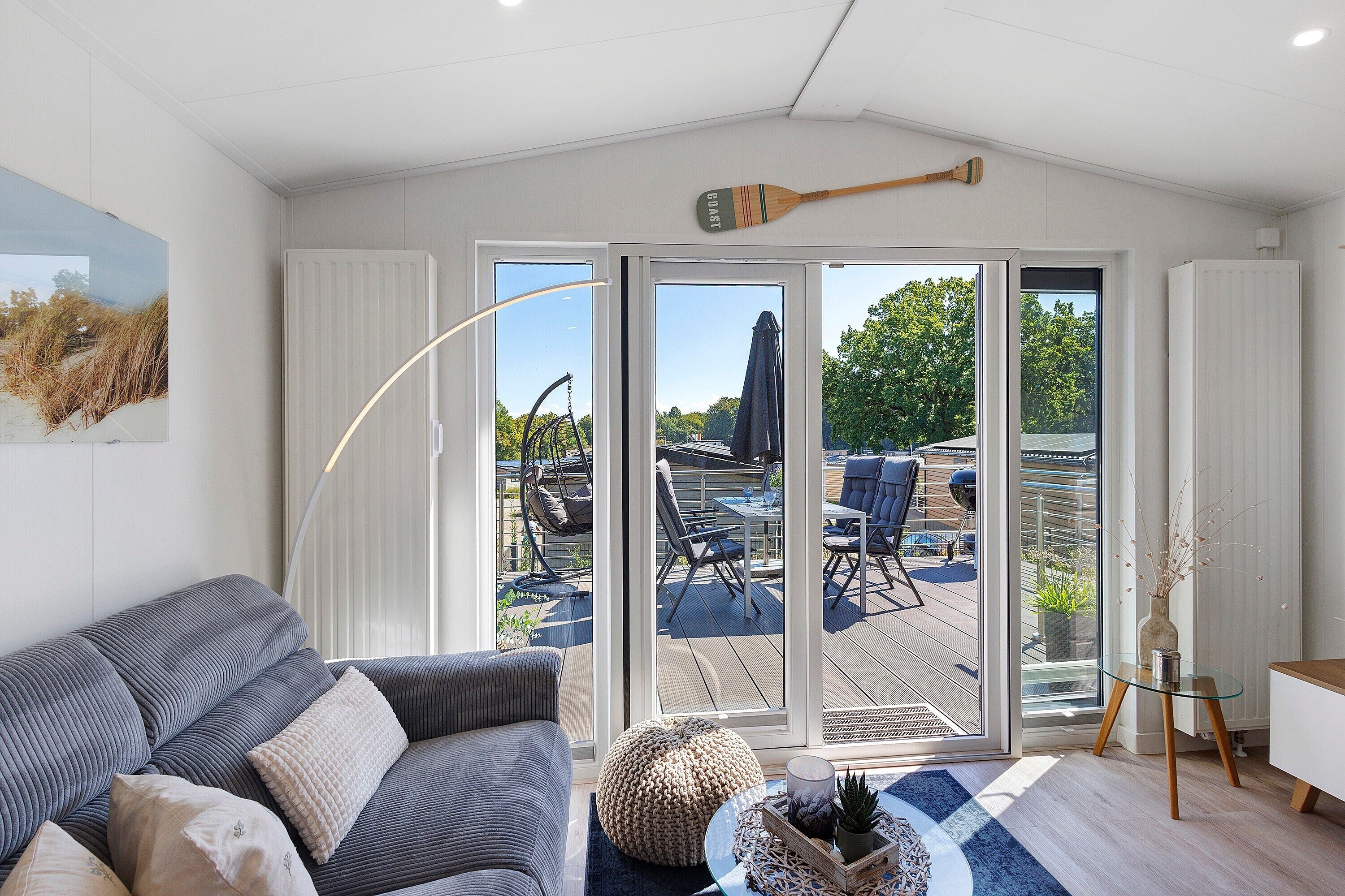 Ein modernes Wohnzimmer mit grauem Sofa, einem Glastisch und Blick auf eine Terrasse mit Gartenmöbeln.