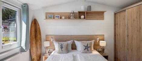 Ein Schlafzimmer mit einem Bett, zwei Nachttischen, einem Holzkopfstück, einem Surfbrett und einem Fenster mit Blick auf Grünflächen.