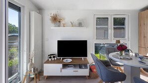 Moderni olohuone, jossa on litteänäyttöinen televisio, ruokapöytä tuoleineen ja suuri ikkuna, josta on näkymä ulos.