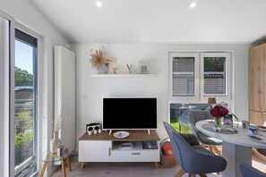 Ein modernes Wohnzimmer mit Flachbildfernseher, einem Esstisch mit Stühlen und einem großen Fenster mit Blick ins Freie.