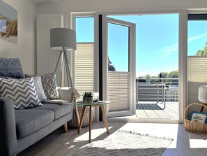 Ein modernes Wohnzimmer mit grauem Sofa, gemusterten Kissen, einer Stehlampe, einem kleinen Tisch mit Pflanze und Blick durch eine Glastür zu einer Terrasse.