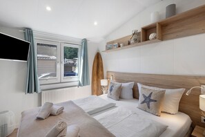 Una habitación de hotel con cama, televisión, ventana con cortinas y estante de madera con objetos decorativos.