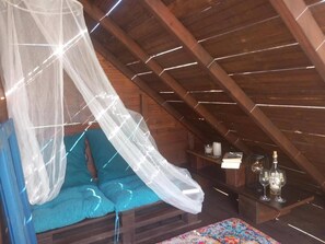 Ein Zimmer mit Holzbalkendecke, einem Bett mit blauer Matratze und einem weißen Baldachin, einem kleinen Tisch mit Kerzen und Gläsern sowie einem farbenfrohen Teppich.