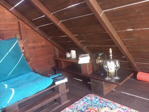 Ein Dachzimmer mit Holzbalkendecke, einem blauen Sitzkissen, einem kleinen Tisch mit Kerze, Laterne und einer Flasche.
