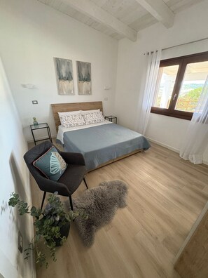 Une chambre à coucher avec un lit, une chaise, une petite table, une plante et une fenêtre avec des rideaux.