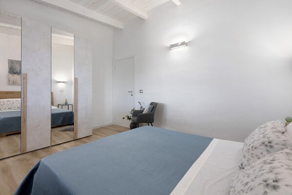 A bedroom with a bed, a chair, a mirror, and a wall-mounted light fixture.
