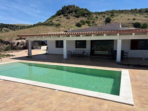 A modern house with a swimming pool, patio seating, and a view of a hilly landscape.
