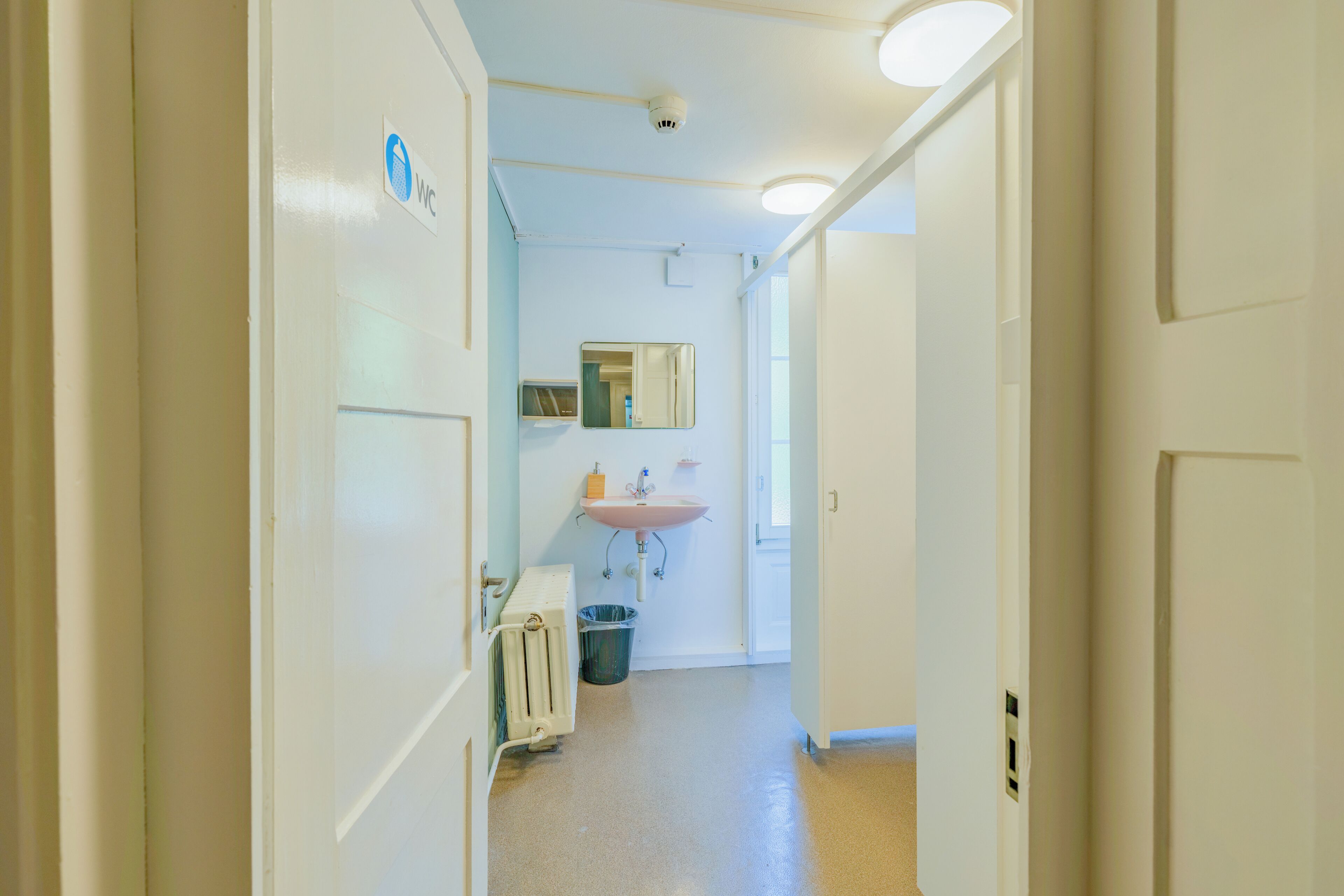 Un pasillo estrecho con una puerta blanca a la izquierda, un radiador blanco, un cubo de basura y un lavamanos con un espejo encima.