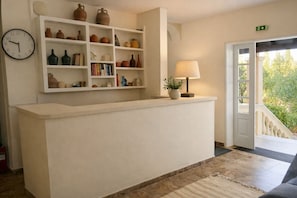 A reception desk with a clock, shelves with pottery and books, a lamp, and a door leading outside.