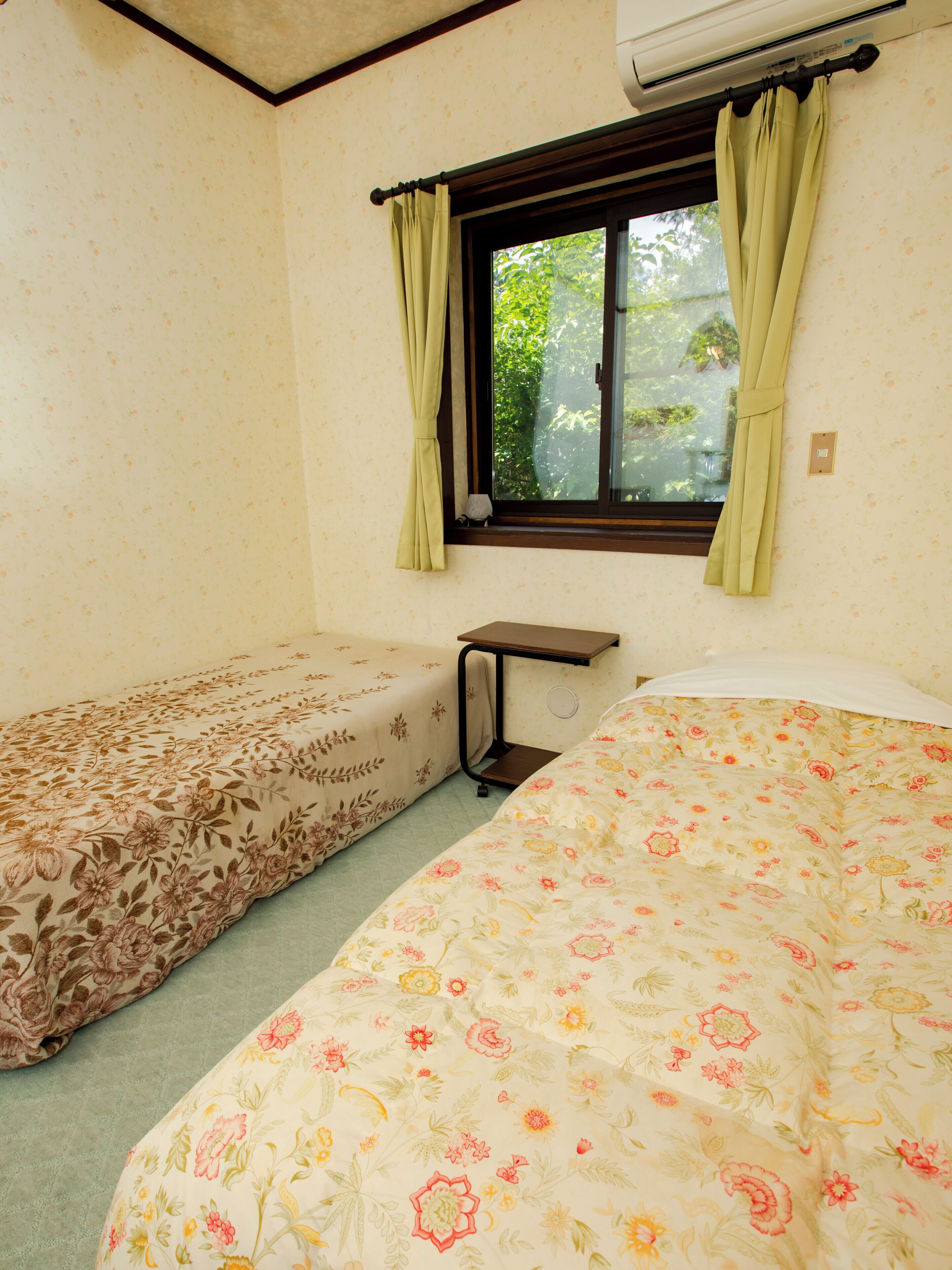 A room with two beds, a window with curtains, a small table, and an air conditioning unit.