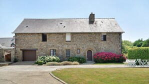 Un edificio in pietra con un ingresso centrale, un piccolo portico e un giardino con fiori e arbusti.