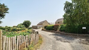 Strada di campagna che conduce a edifici in pietra, con una recinzione in legno e vegetazione.