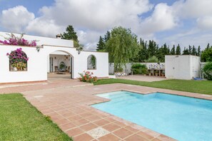 Ein Pool mit Terrassenbereich, ein weißes Gebäude mit Bogenportal und ein Garten mit Bäumen und Blumen.