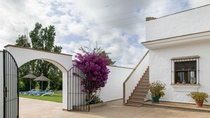Un cancello con una scalinata che conduce a un edificio bianco, una pianta fiorita viola e vista su un prato con poltrone da relax.