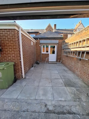 Une petite cour étroite, avec un mur en briques, une poubelle verte et une porte donnant accès à un bâtiment.