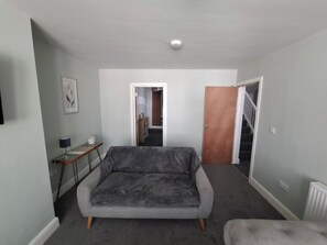 A room with a grey sofa, a small table with a lamp, a framed picture on the wall, and a door leading to another room.