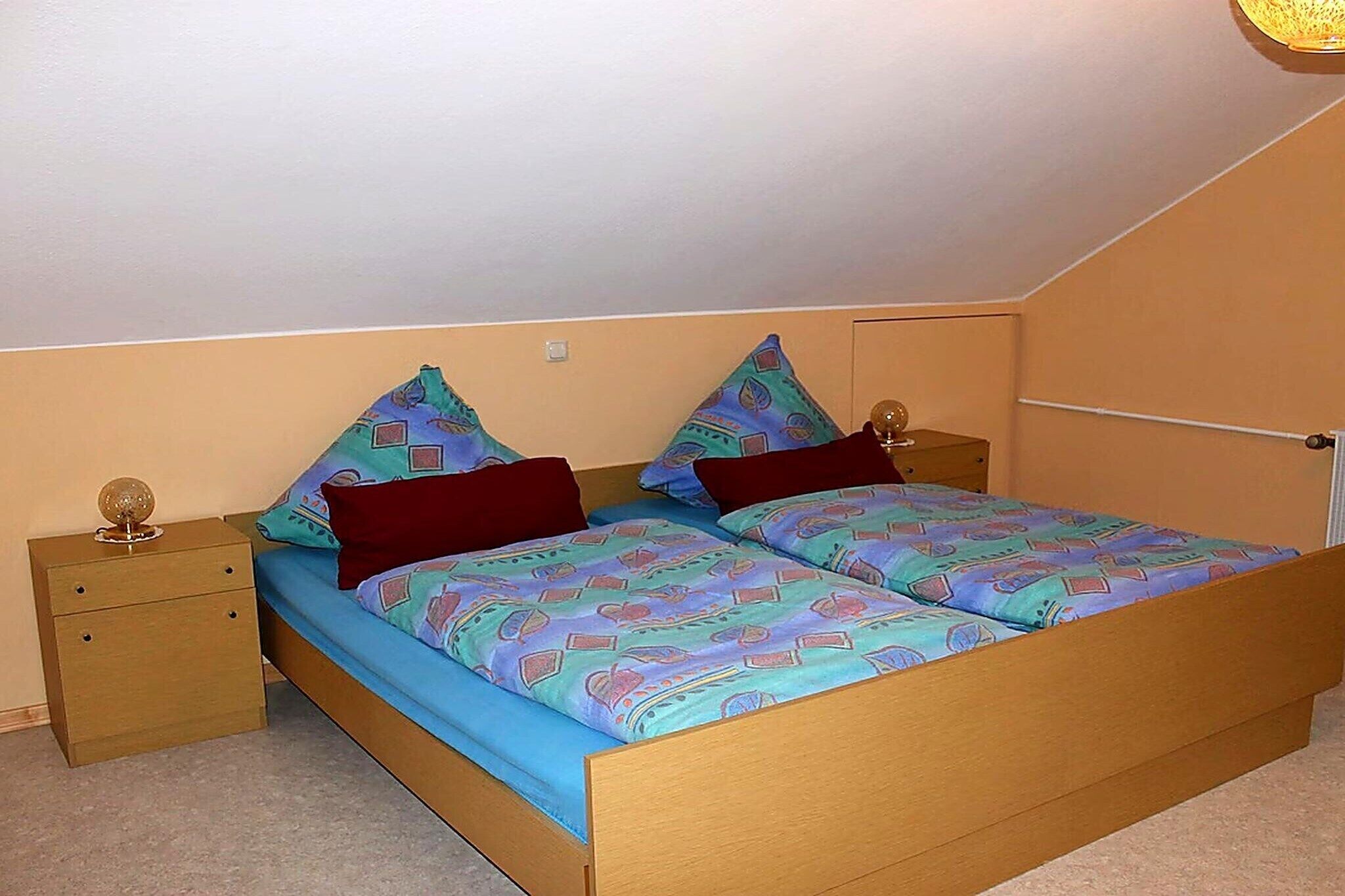 A single bed with a patterned bedspread, two pillows, a nightstand with a lamp, and a wall-mounted shelf.