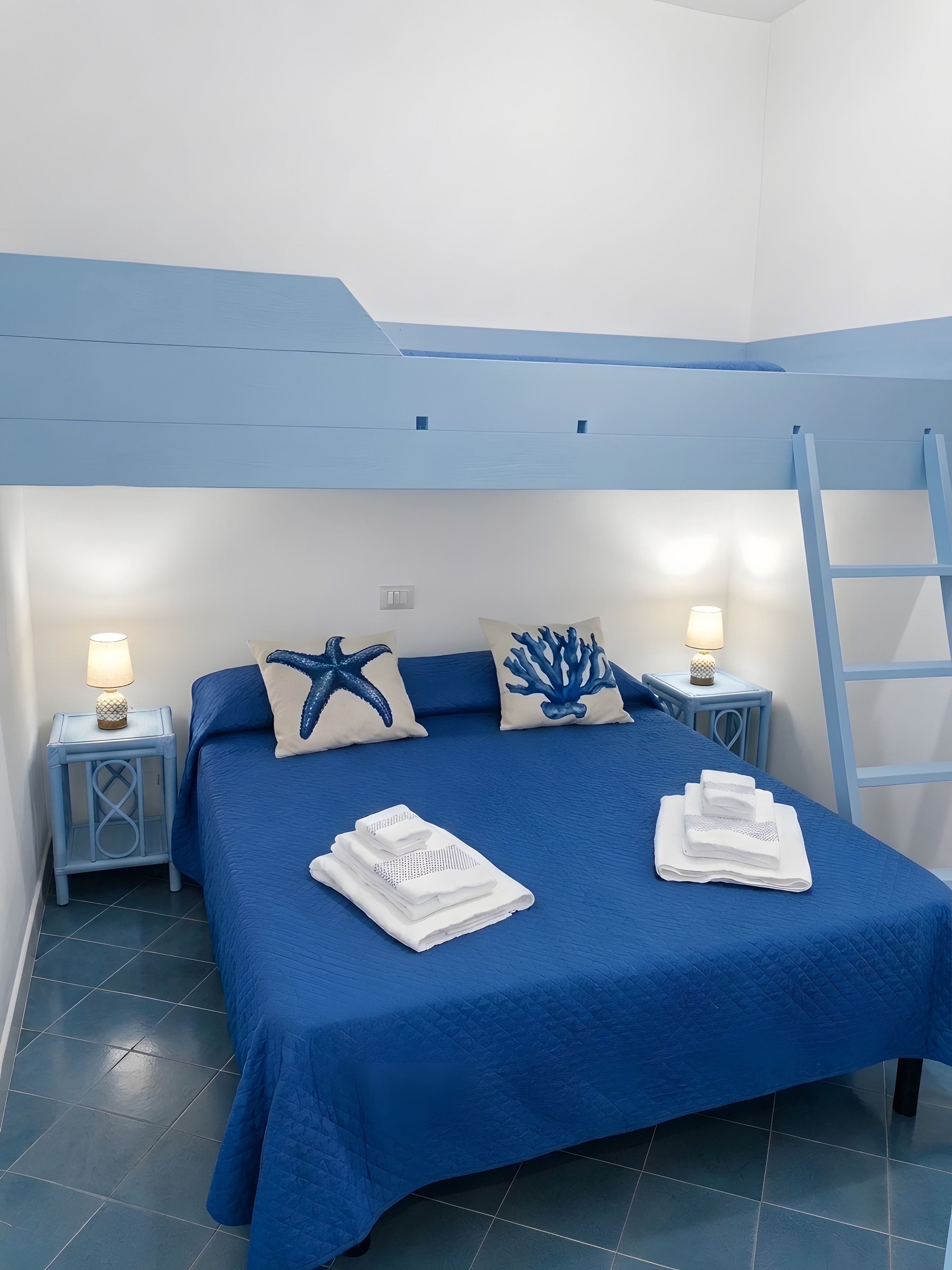 Un dormitorio con una cama litera, dos mesitas de noche con lámparas, una almohada con forma de estrella de mar, una almohada con forma de coral y toallas dobladas sobre la cama.