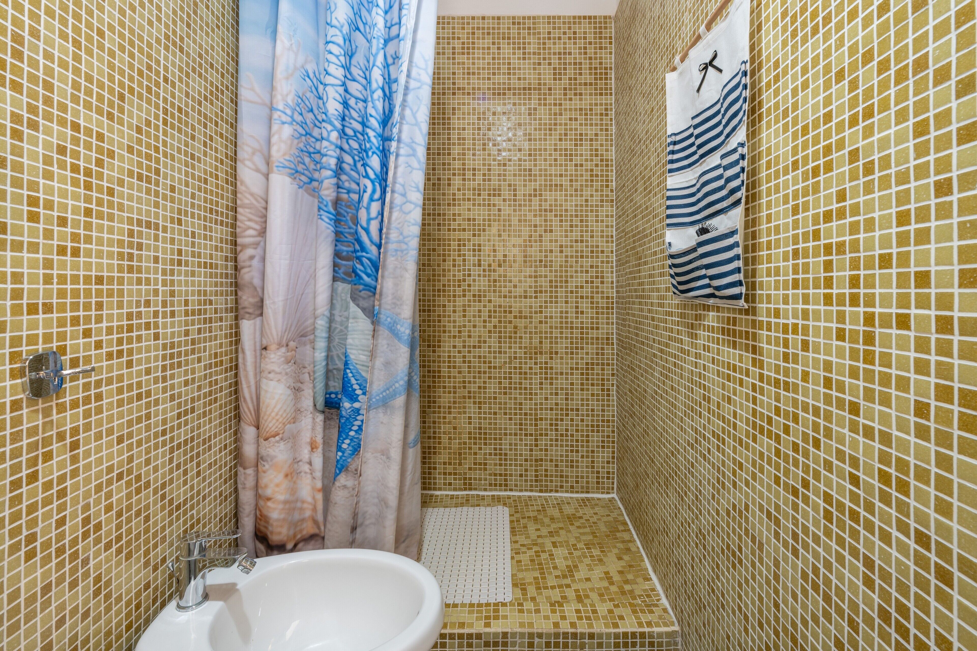 Un bagno con pareti in mosaico, una tenda da doccia con motivo floreale, un lavandino bianco e un portasciugamani con asciugamani a righe.