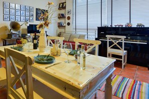 A dining area with a wooden table, chairs, a vase with flowers, a piano, and a window with blinds. - Holiday Home 'Wine & Piano Stories' with Private Terrace, Shared Garden and Wi-Fi (Esperii)