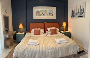 Habitación de hotel con una cama, dos mesitas de noche, un tocador, un espejo, un cuadro abstracto enmarcado y una pintura de un velero.