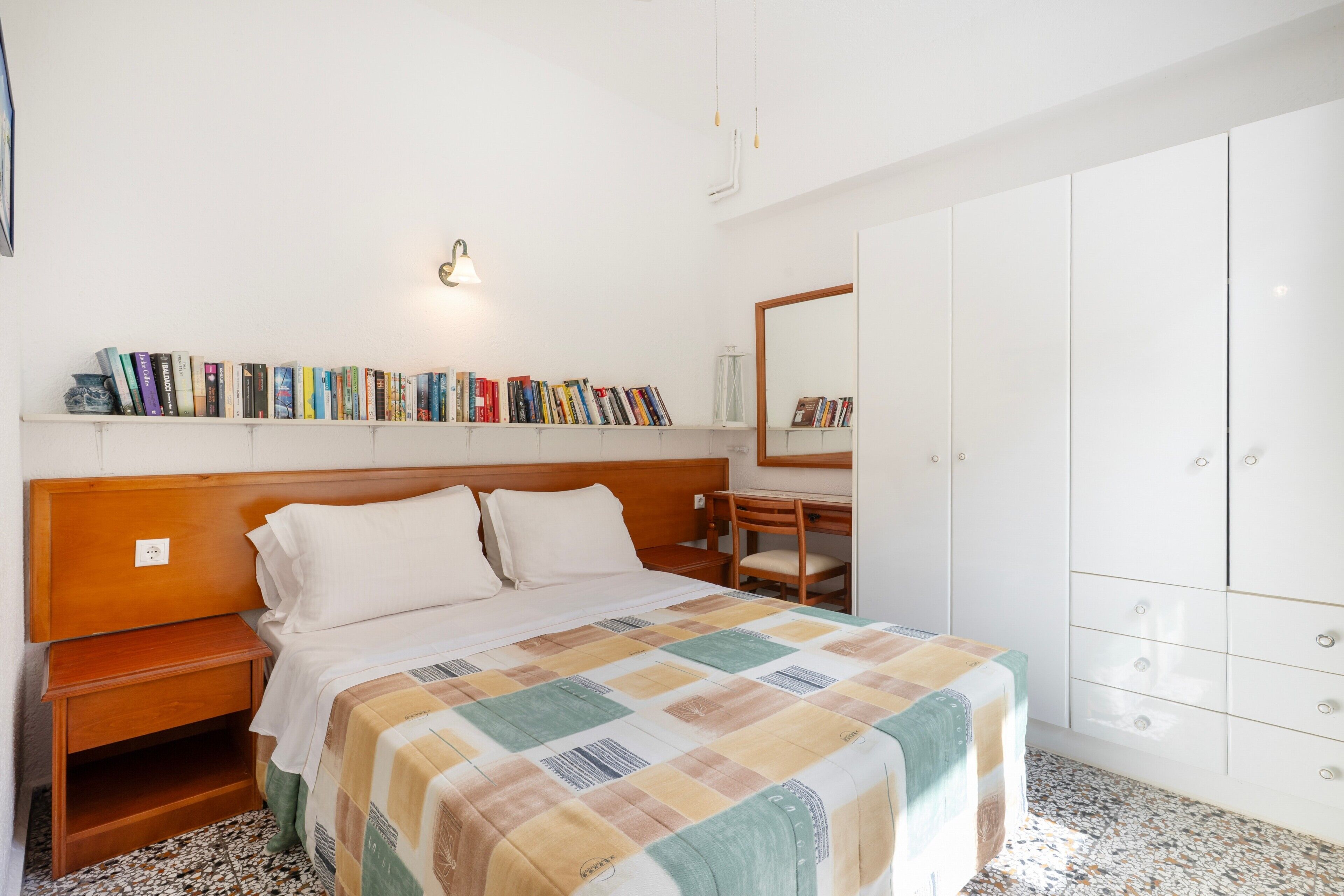 A bedroom with a bed, a bedside table, a chair, a wardrobe, and a shelf with books.