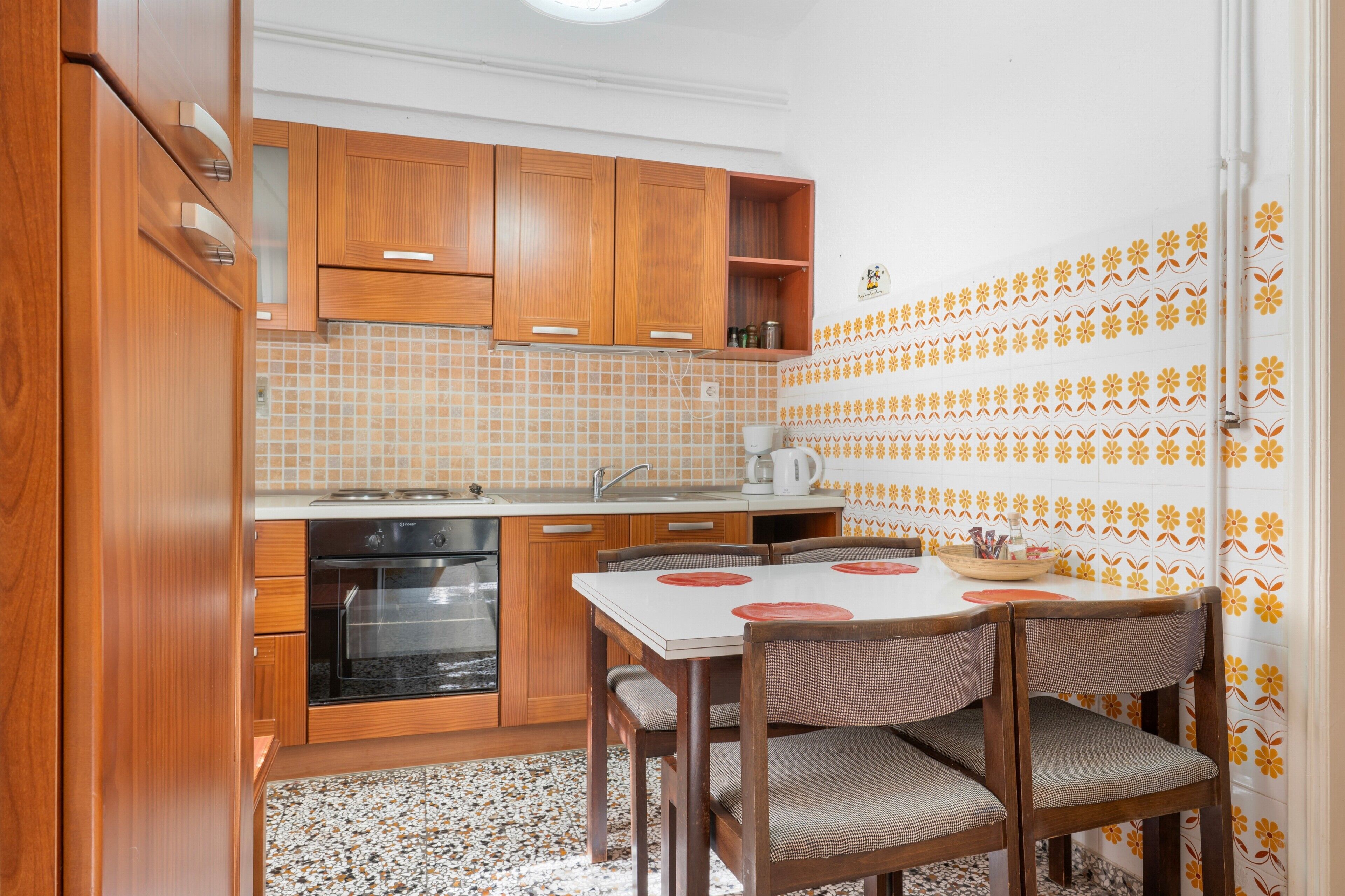 Een compacte keuken met houten kastjes, een inbouwoven, een spoelbak en een eethoek met een tafel en stoelen.