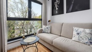 A modern living room with a sofa, a round table, and a large window with a view of trees and buildings. - ShortStayPoland Zgierska B106 (Warszawa)