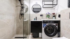 A modern bathroom with a glass shower enclosure, a built-in washing machine, a round mirror, and a small sink area. - ShortStayPoland Zgierska B106 (Warszawa)