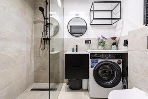 A modern bathroom with a glass shower enclosure, a built-in washing machine, a round mirror, and a small sink area. - ShortStayPoland Zgierska B106 (Warszawa)