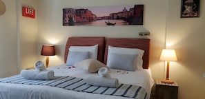 A hotel room with a bed, two bedside lamps, a wall clock, a life sign, and a cityscape painting. - 2BR Gouvia – Kitchenette 6 (Kontokali)