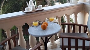 Ein Balkon mit Tisch, gedeckt für das Frühstück, darunter eine Karaffe, Tassen und Gläser Orangensaft. Man sieht Palmen im Hintergrund.