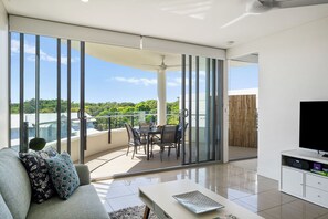 Ein modernes Wohnzimmer mit einer Couch, einem Flachbildfernseher, einer Essecke mit Tisch und Stühlen sowie einem Balkon mit Blick auf Grünflächen.