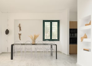 Ein Essbereich mit Tisch, Stühlen, einer Vase mit getrockneten Pflanzen und einem Fenster mit grünem Rahmen.