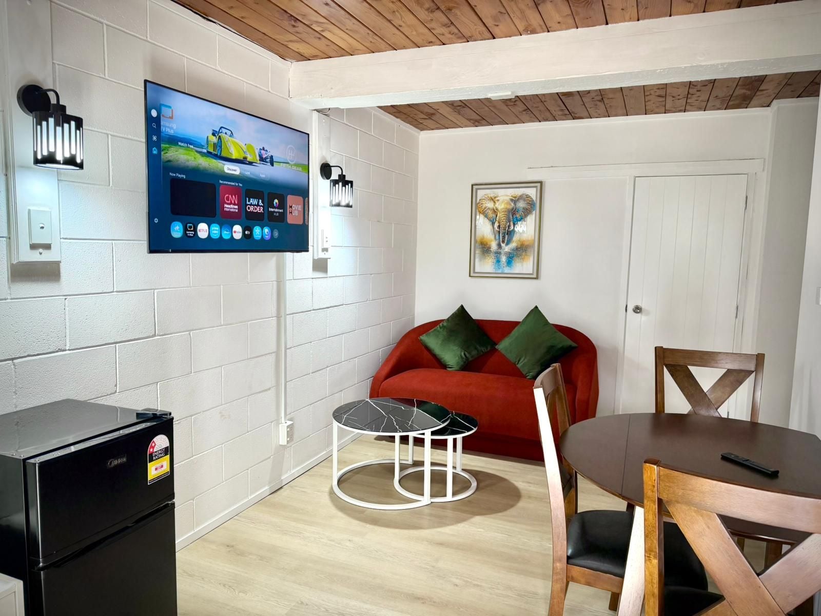 A modern living room with a flat-screen TV mounted on the wall, a red sofa with green cushions, a round wooden dining table with chairs, and a black refrigerator.