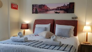 A hotel room with a bed, two bedside lamps, a wall clock, a life sign, and a cityscape painting. - Family 2BR Apt Pool – Kitchenette 7 (Kontokali)