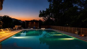 A swimming pool with a wooden deck and lounge chairs, surrounded by trees and a building with lights. - Family 2BR Apt Pool – Kitchenette 7 (Kontokali)